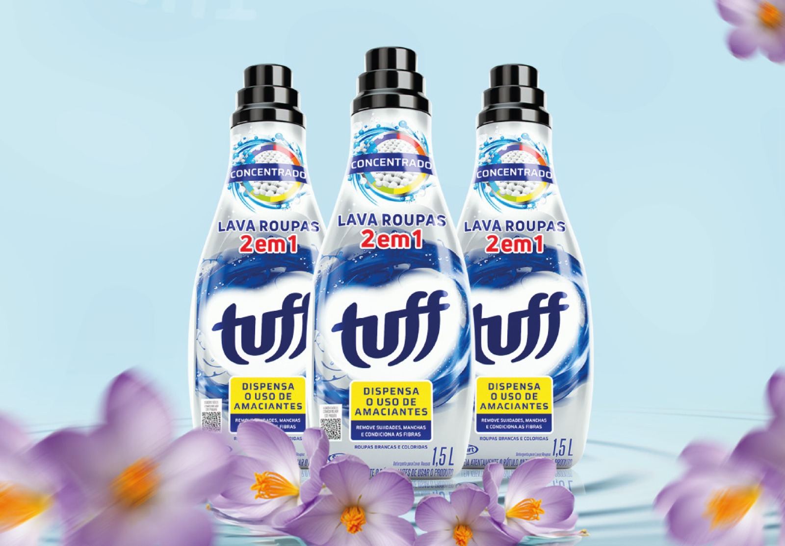 Três frascos de lava-roupas concentrado 2 em 1 Tuff de 1,5L alinhados sobre fundo azul claro, com flores roxas desfocadas em primeiro plano.