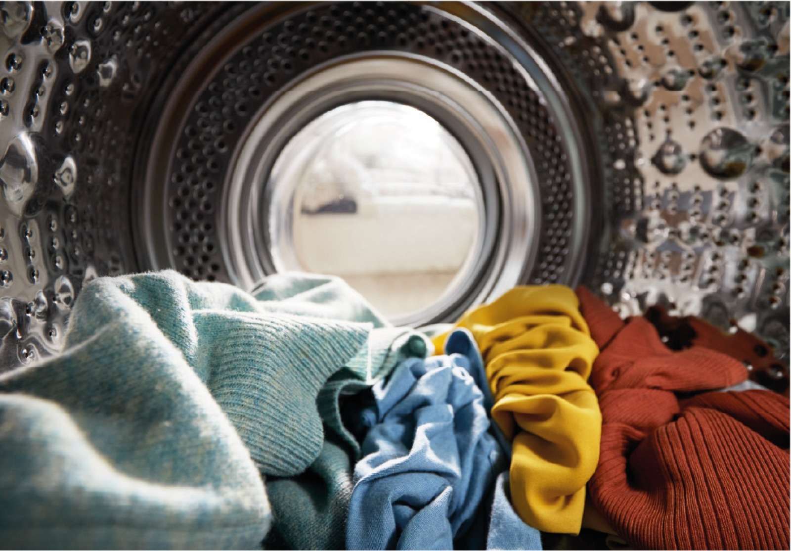Visão interna do tambor de inox de uma máquina de lavar roupas, com peças coloridas em tons de azul, amarelo e terracota no fundo. Visão interna do tambor de inox de uma máquina de lavar roupas, com peças coloridas em tons de azul, amarelo e terracota no fundo.